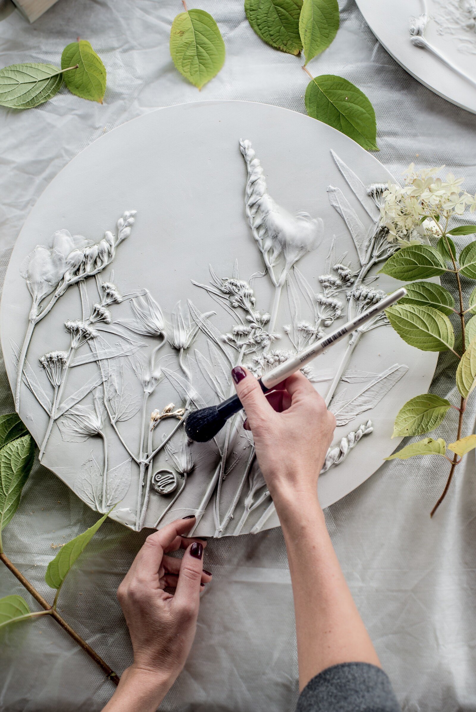 Hands brushing a round plaster plaque with botanical relief, surrounded by green leaves on fabric