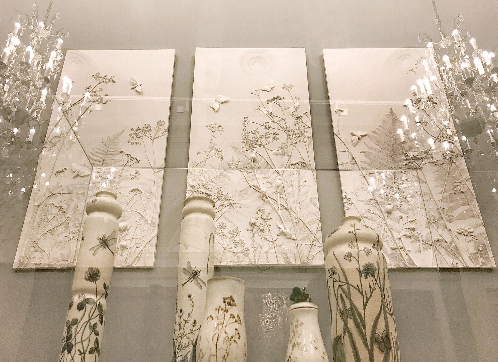 Three white botanical relief panels with chandeliers and decorated vases displayed in a window