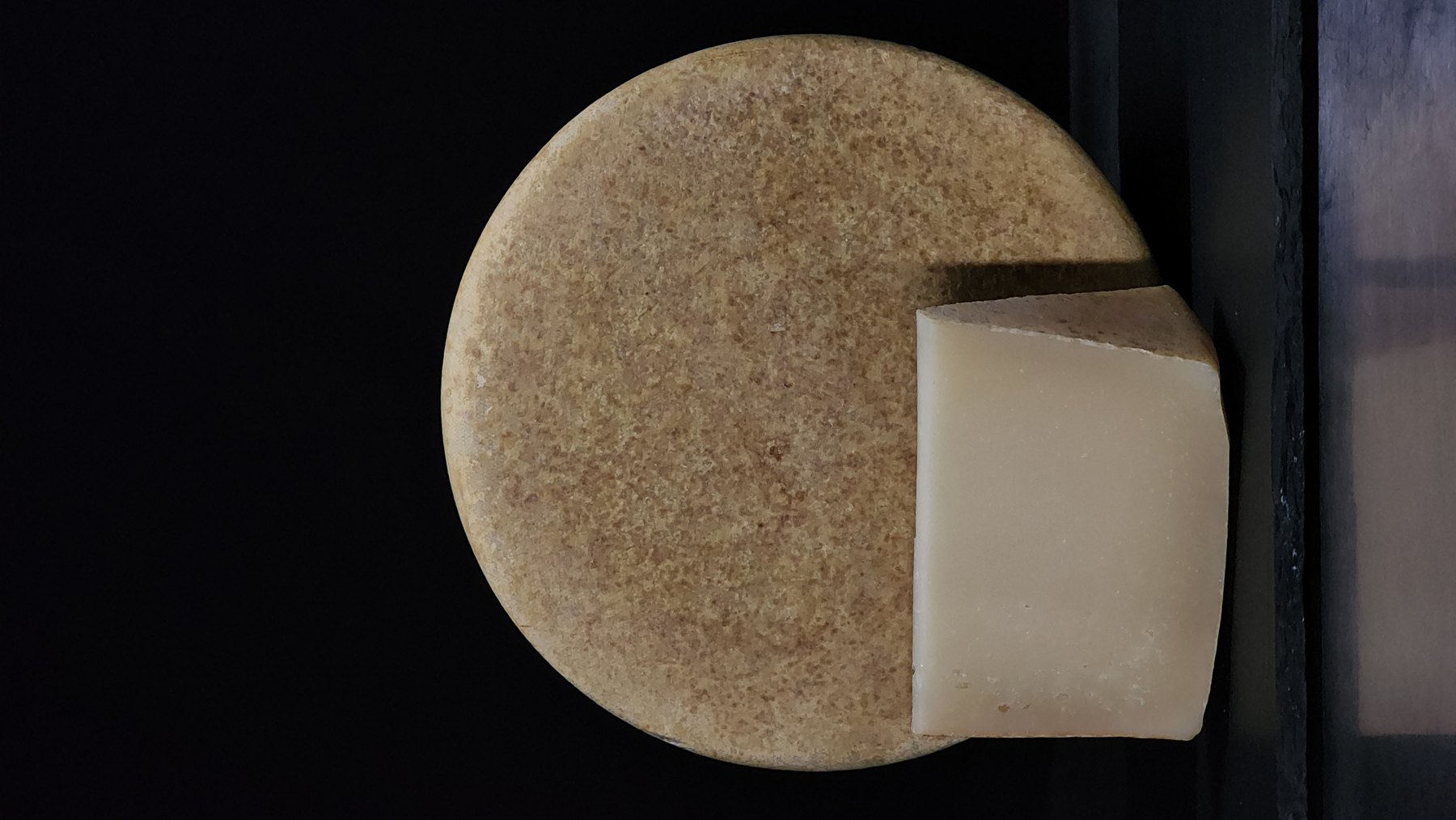 Round pale cheese wheel with a cut wedge beside it on a dark background