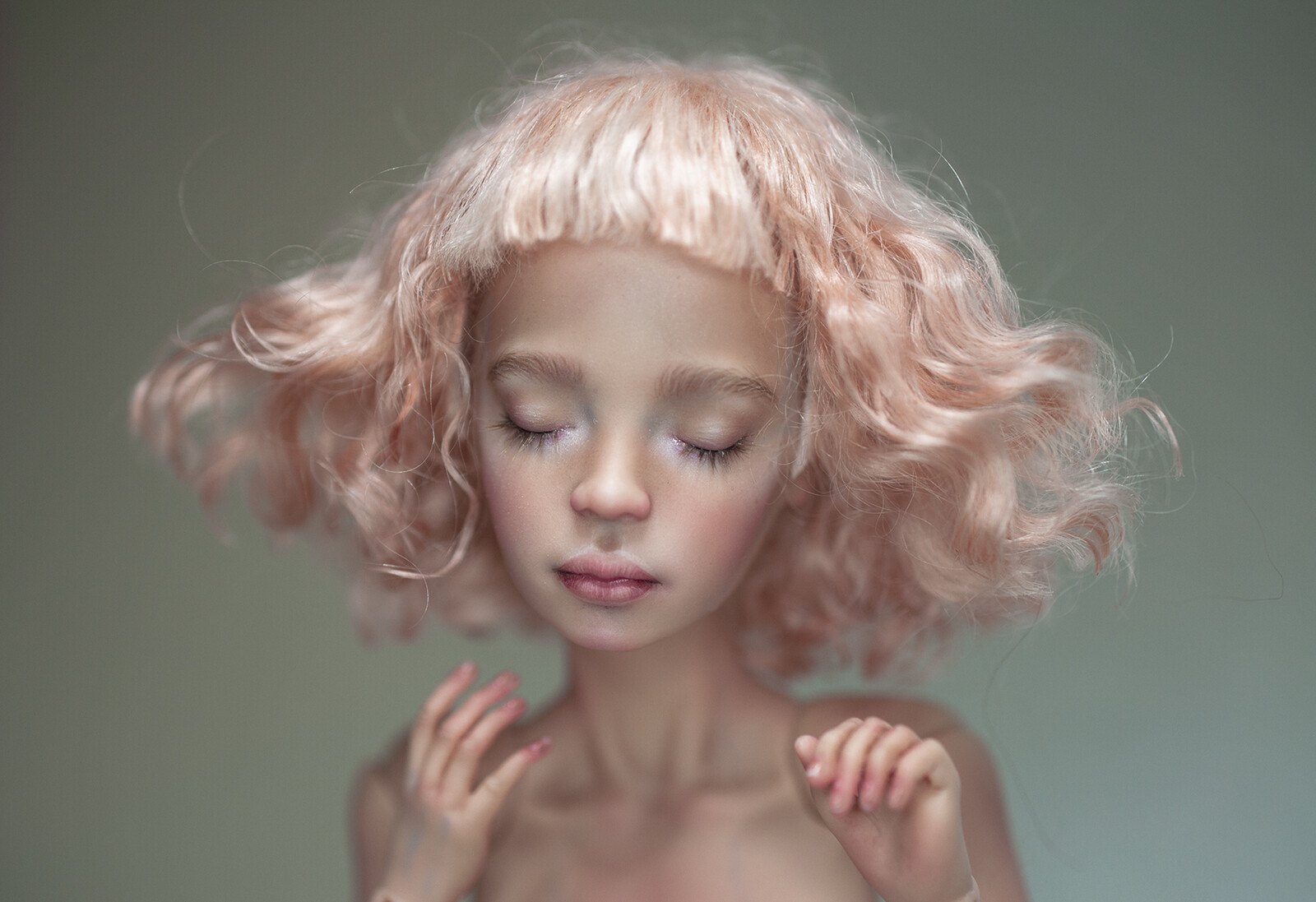 Doll with closed eyes and curly peach-pink hair, hands raised near the shoulders