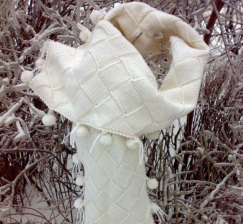 A cream entrelac knitted scarf with pom-poms and fringe hanging on snowy branches