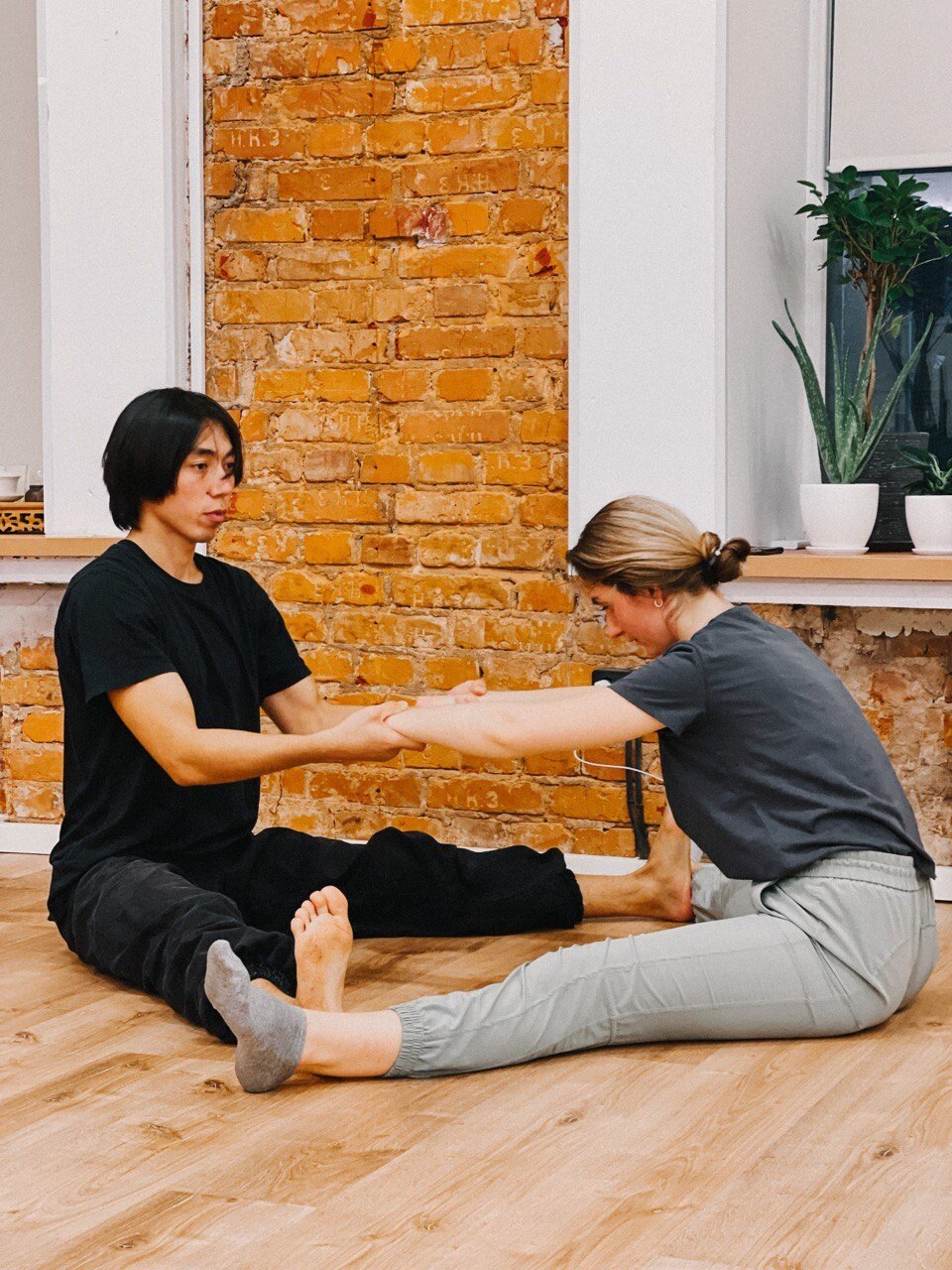 Instructor helping a woman in a seated forward stretch in front of a brick wall