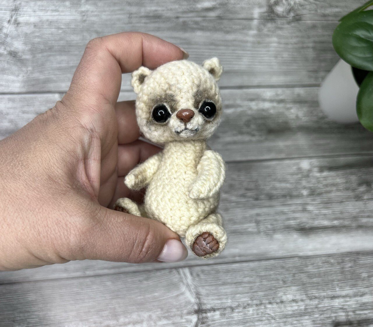 A hand holding a small cream crocheted bear with black eyes against gray wood