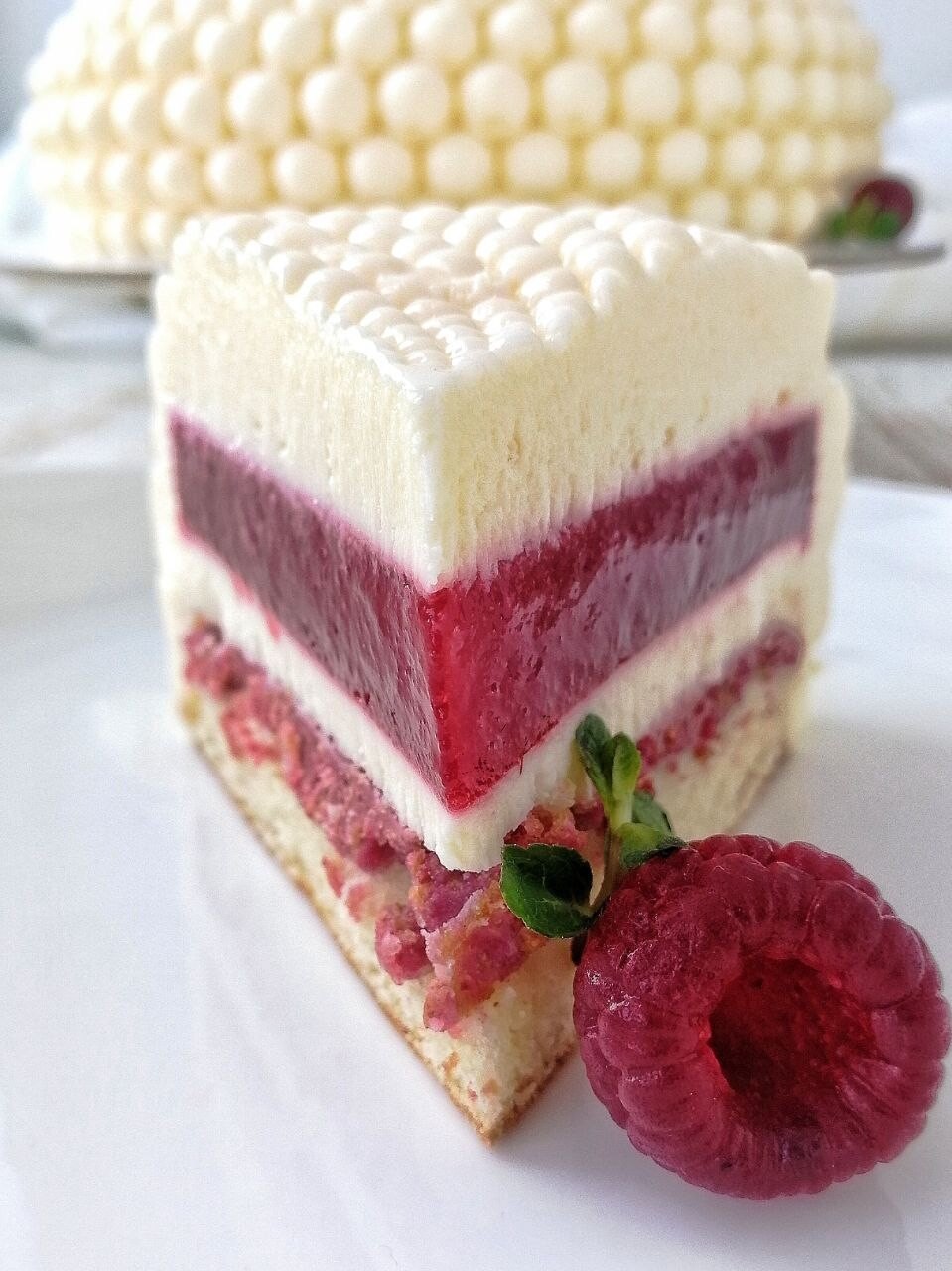 Slice of layered raspberry and white mousse cake with a fresh raspberry beside it