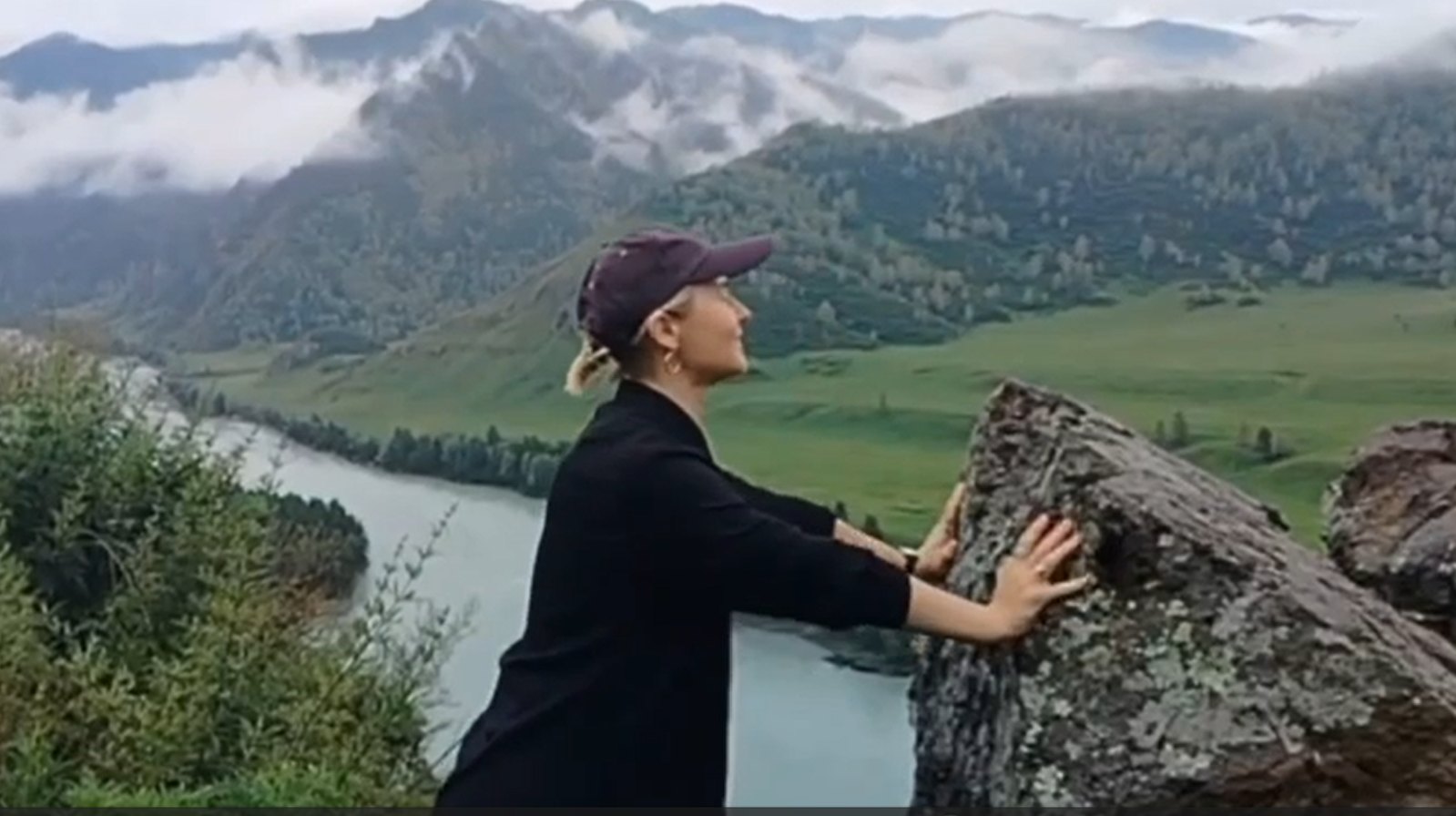 Woman in a cap touching a mossy rock above a misty river valley