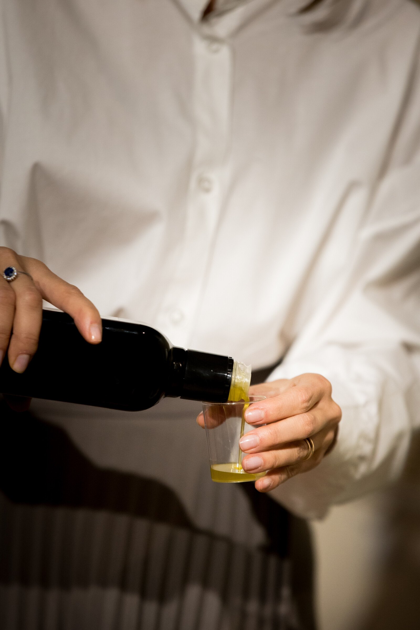 Person in a white shirt pouring olive oil from a dark bottle into a small tasting cup