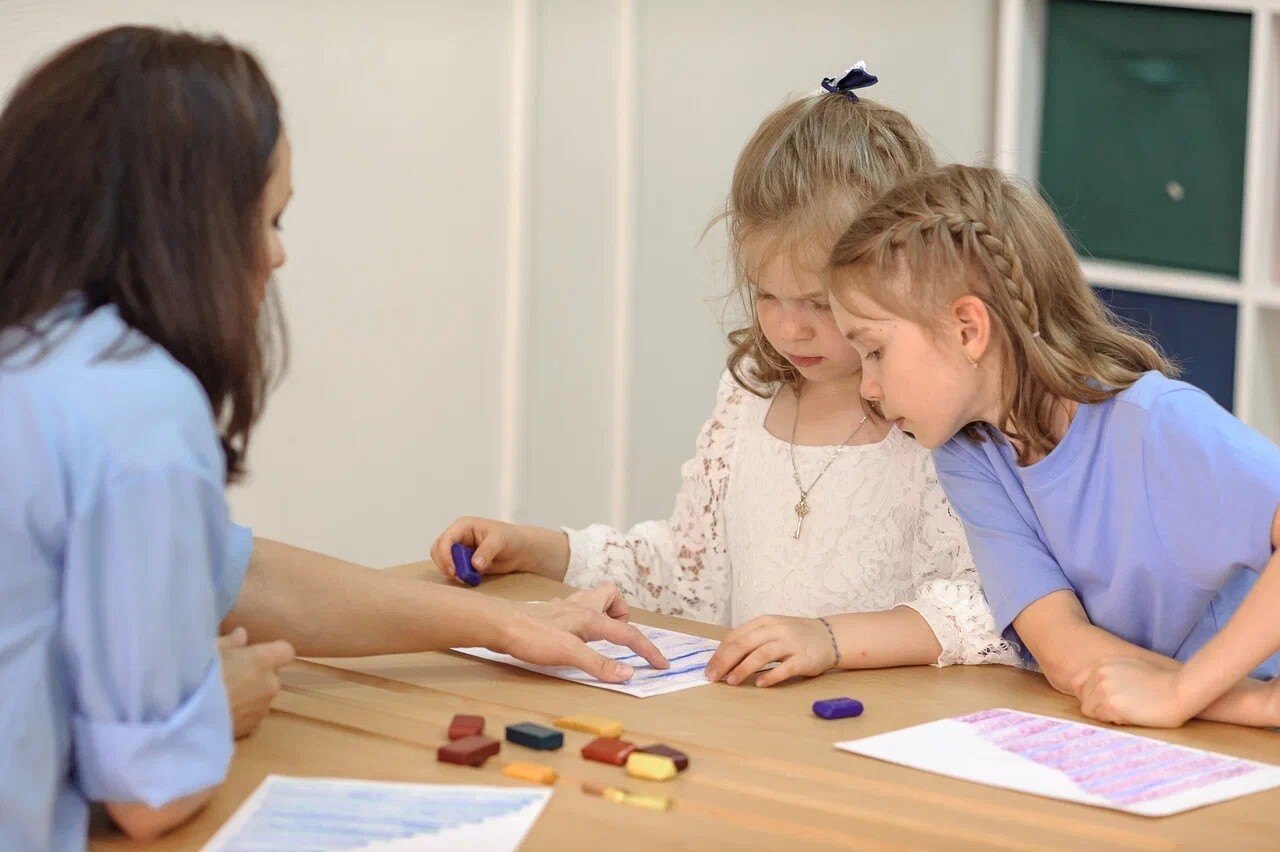 Woman and two girls drawing with wax blocks on paper at a wooden table