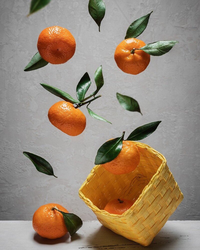 Tangerines with leaves falling into a tilted woven basket against a gray wall