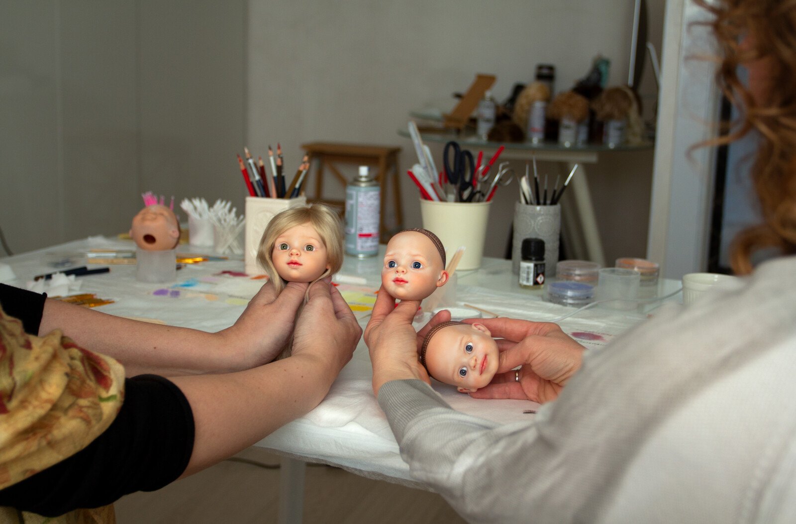 Hands holding three painted doll heads above a workshop table with brushes and supplies