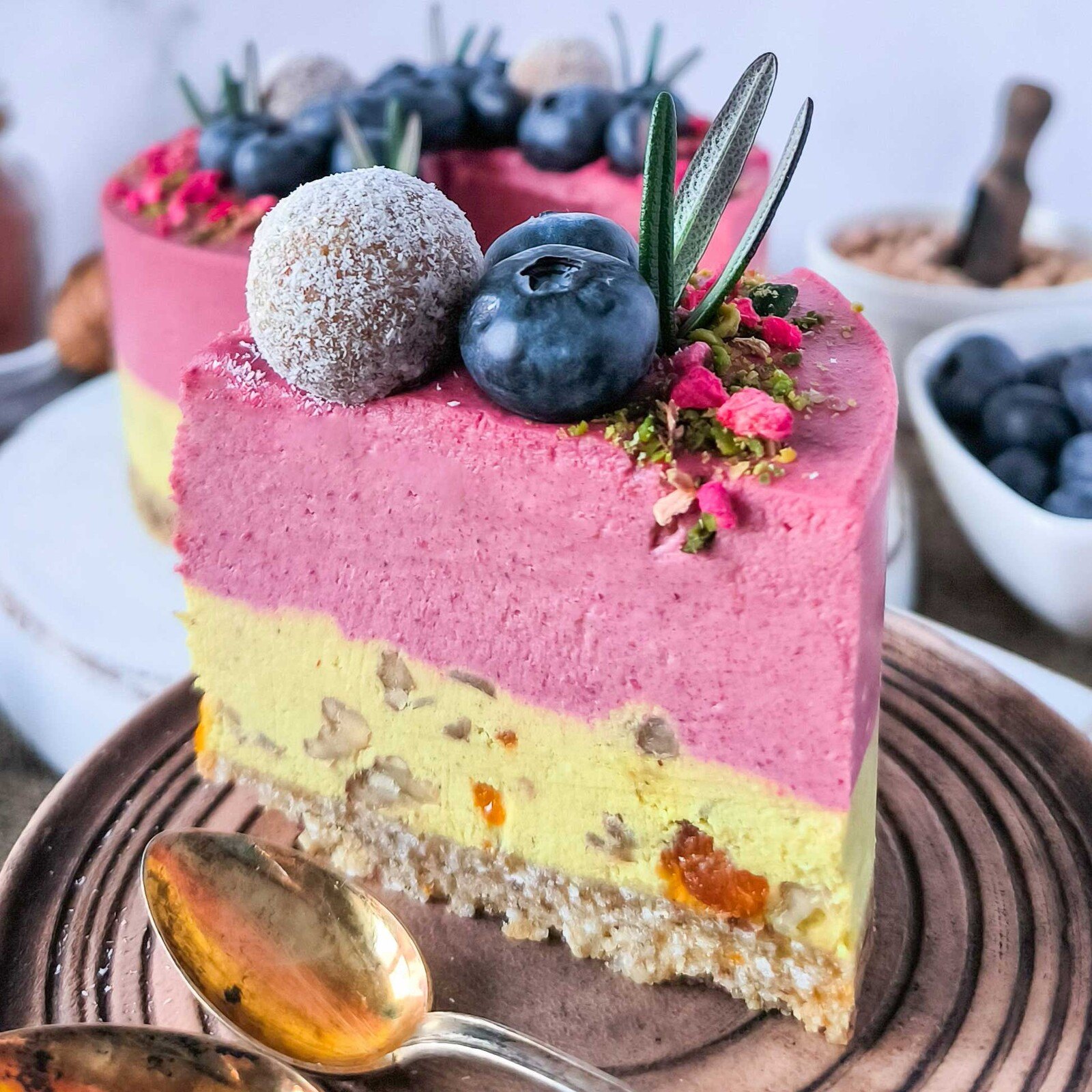 Slice of pink and yellow raw cake with blueberries, a coconut ball and rosemary