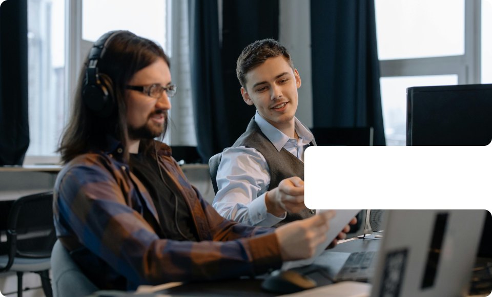 Two coworkers in an office discussing a printed document, one wearing headphones and glasses