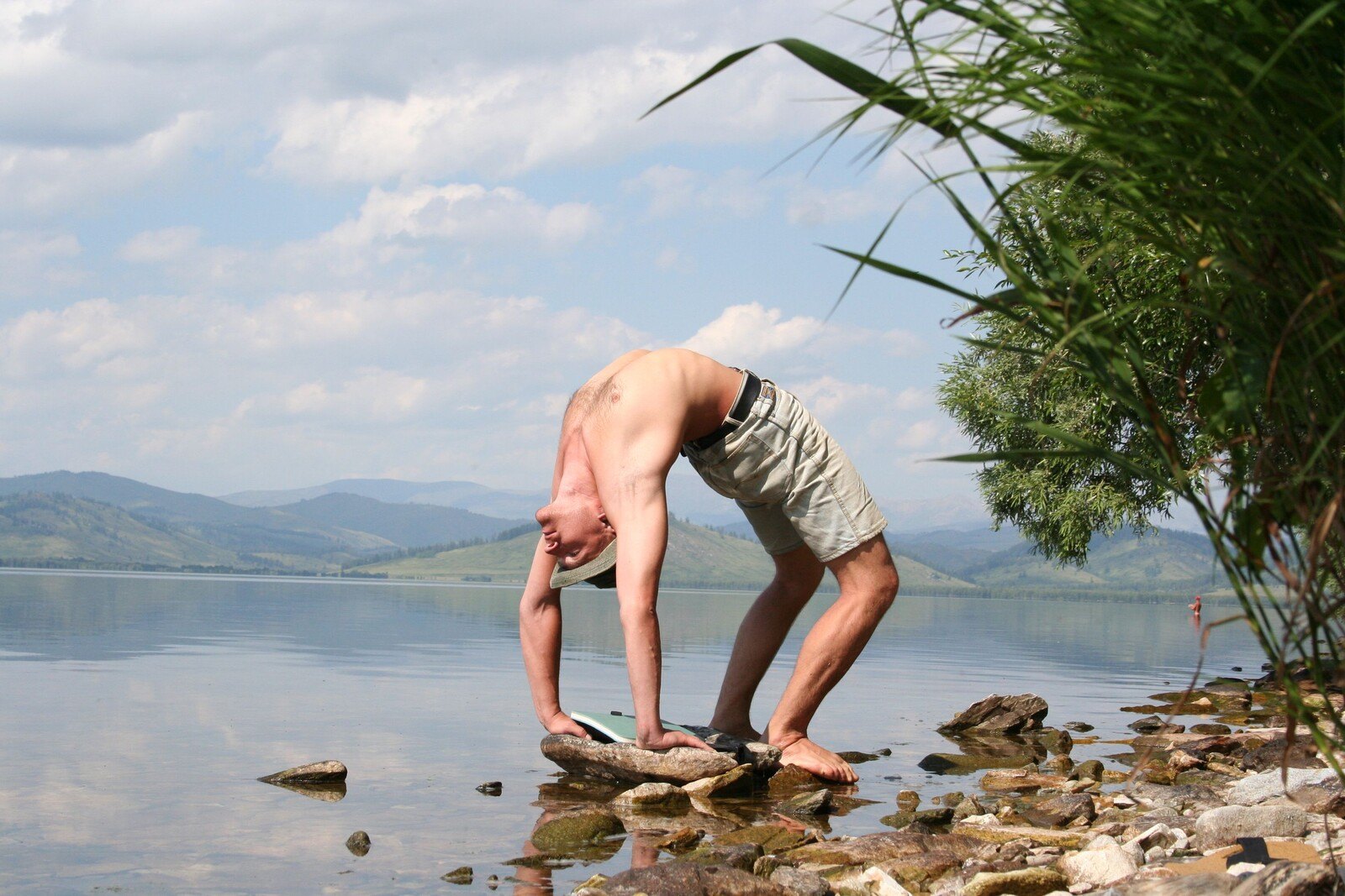 Man performing a standing backbend on a rocky lake shore with mountains behind