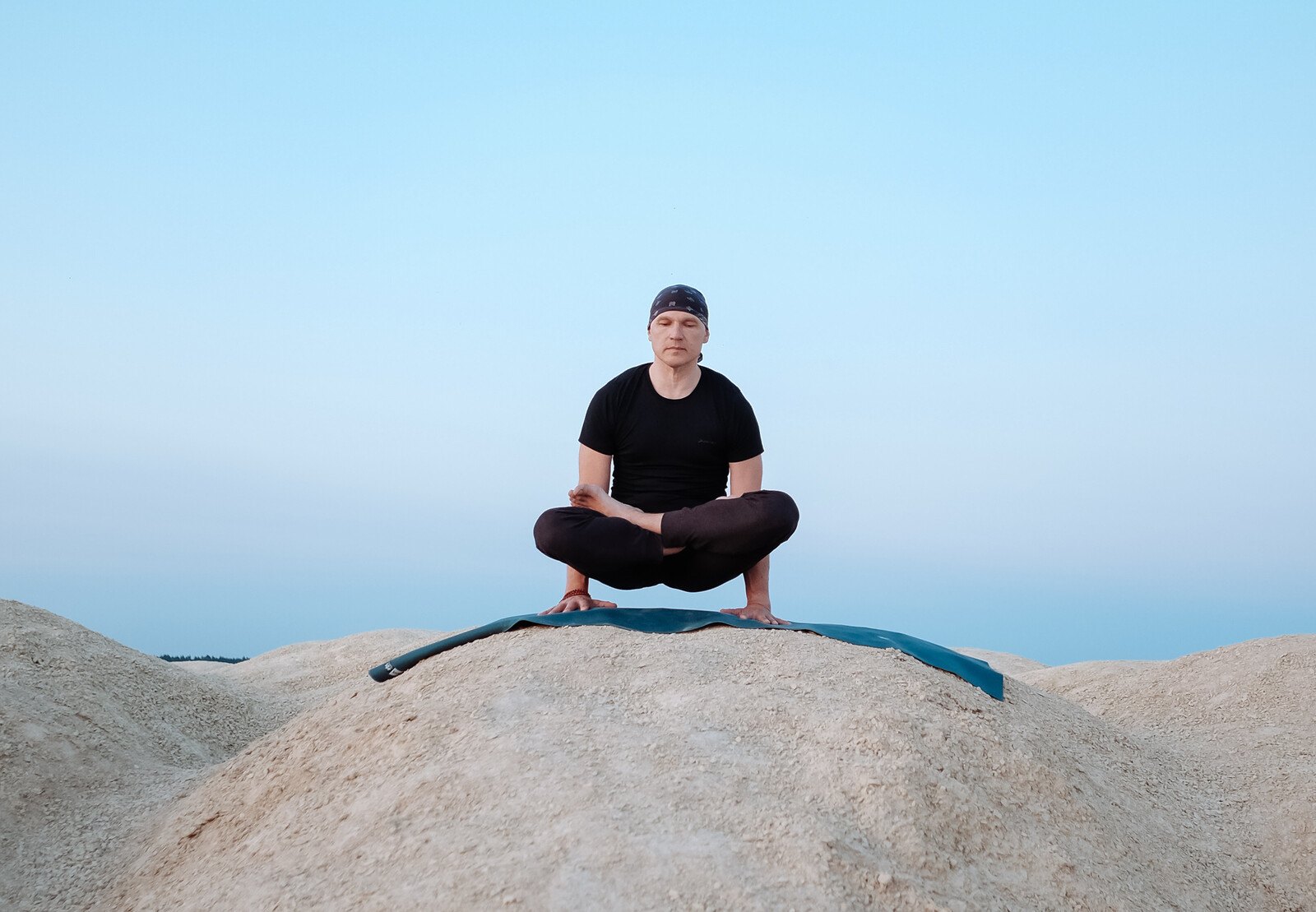 Man balancing in an arm-balance yoga pose on a mat on a sandy hill